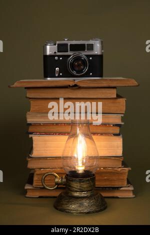 Libri antichi e una lampada a mano con lampadina, stilizzata come antica. C'è una vecchia macchina fotografica in cima ai libri. Oggetti collocati su uno sfondo di oliva. Foto Stock