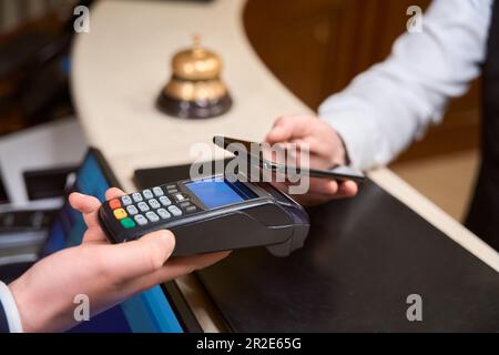 Uomo che effettua il pagamento con telefono, receptionist con lettore di carte di credito in hotel Foto Stock