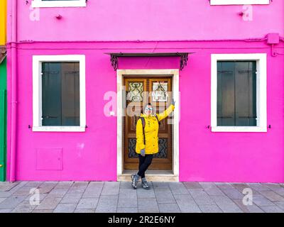Donna sorridente in posa di fronte a casa rosa a Burano Island. Felice viaggiatore turista femmina con impermeabile giallo e occhiali da sole in posa e divertirsi Foto Stock