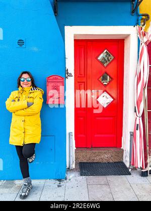 Felice viaggiatore donna turista con impermeabile giallo e occhiali da sole posa all'ingresso della casa blu con porta rossa, casella postale sull'isola di Burano, Venezia Foto Stock