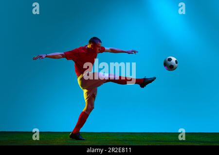 Ritratto di calcio, giocatore di calcio in uniforme sportiva calci la palla in movimento su sfondo campo in luce al neon. Colpire la palla con la gamba Foto Stock