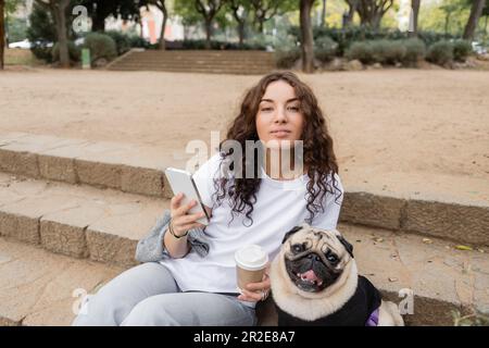 Giovane donna sorridente ricci in abiti casual utilizzando il telefono cellulare e tenendo il caffè per andare mentre si guarda la macchina fotografica vicino al cane pug seduto sulle scale in sfocatura Foto Stock