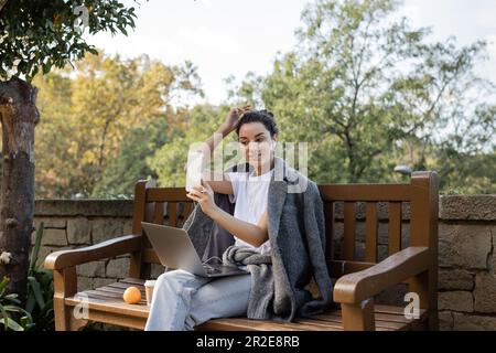 Giovane e sorridente freelancer in giacca calda con auricolari e smartphone mentre si siede vicino a un computer portatile, drink da asporto e arancione fresco sulla panca di legno Foto Stock