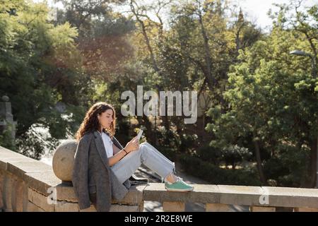 Donna giovane e ricciola in giacca calda e abiti casual con smartphone e auricolari con cavo mentre si siede sulla recinzione e si passa del tempo in un parco sfocato Foto Stock