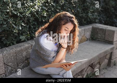 Donna giovane e ricciola in abiti casual che toccano le labbra, tenendo l'indicatore e guardando il notebook mentre si siede su una panca di pietra vicino a cespugli verdi in Barcel Foto Stock