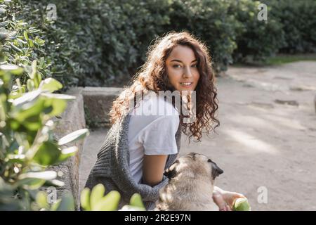 Giovane donna curly spensierata in t-shirt e maglione tenendo la mela verde e guardando la macchina fotografica mentre si siede su una panca di pietra vicino al cane pug e rilassarsi dentro Foto Stock