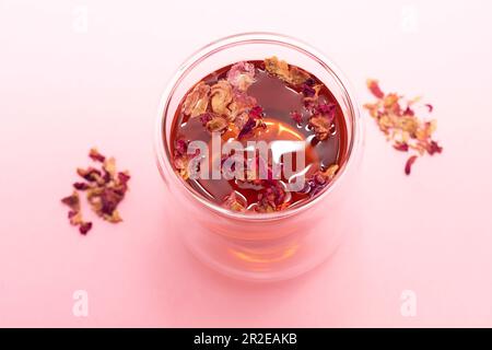 Sani petali di rosa alle erbe, boccioli di tè in tazza di vetro a doppia parete su sfondo rosa. Bevanda a base di erbe essiccata di petali di fiori colorati, bevanda Foto Stock