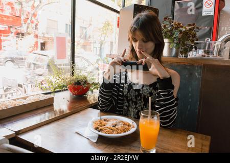 vista frontale della giovane donna transgender latina di etnia argentina, scattando una foto del suo cibo con il telefono, seduto nel ristorante, ristorante Foto Stock