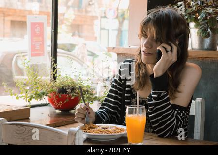 giovane transgender donna latina di etnia argentina, seduta a parlare al telefono mentre si mangia pasta in un ristorante, stile di vita concetto, copia spazio Foto Stock