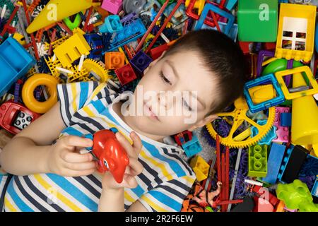 Simpatico ragazzino che gioca con i giocattoli che giacciono tra i giocattoli colorati sul pavimento, vista dall'alto Foto Stock