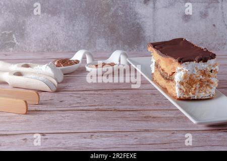 Primo piano di una milla-feuille farcita di crema e mandorle ricoperte di cioccolato su un piatto bianco con cucchiai di ceramica con cioccolato e cacao in polvere Foto Stock