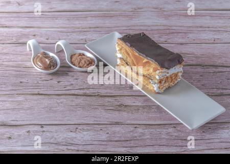Primo piano di una milla-feuille farcita di crema e mandorle ricoperte di cioccolato su un piatto bianco con cucchiai di ceramica con cioccolato e cacao in polvere Foto Stock