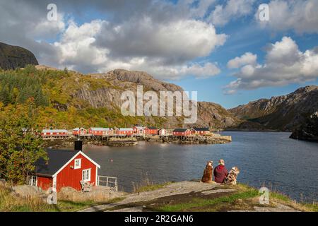 Donna seduta con cani sulla riva. Guardando le capanne rosse dal fiordo, Nusfjord, Flakstad, Lofoten, Norvegia. Foto Stock