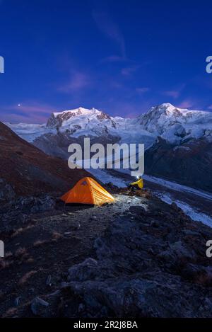 Escursionista con lampada frontale seduto accanto alla tenda di fronte al massiccio del Monte Rosa, al ghiacciaio Gorner (Gornergletscher) e alle cime del Lyskamm al crepuscolo Foto Stock