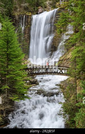 Due persone a piedi attraverso il ponte con la cascata Cirque de Saint-Meme in background, Hauts de Chartreuse Riserva Naturale, Chartreuse, Vercors, SA Foto Stock