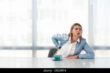 Giovane donna d'affari che ha mal di schiena da cattiva postura mentre seduto alla scrivania in ufficio su sedia non ergonomica al posto di lavoro in abbigliamento formale wi Foto Stock