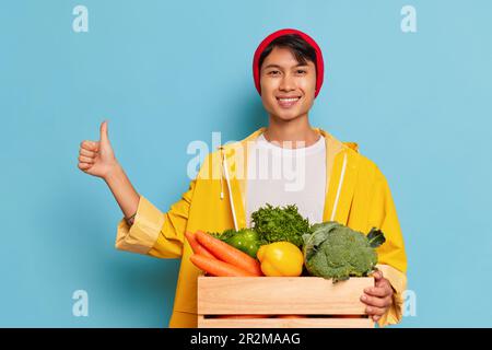 Greengrocer maschile asiatico con giacca gialla e cappello rosso in posa con scatola di verdure mostra con il pollice in su, si trova su sfondo blu, cibo biologico Foto Stock