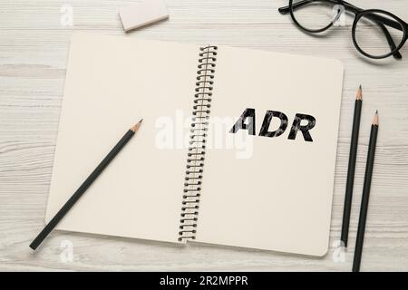 Soluzione alternativa delle controversie. Taccuino con abbreviazione ADR, matite, gomma e bicchieri su tavolo di legno bianco, piatto Foto Stock