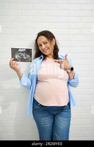 La donna incinta sorride alla macchina fotografica e punta verso l'ecografia al suo bambino, isolato contro una parete di mattoni bianchi. Foto Stock