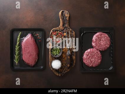 Bistecca di filetto di manzo fresco crudo e hamburger di carne macinata sigillati in vassoio sottovuoto su fondo scuro con spezie e forchetta. Foto Stock