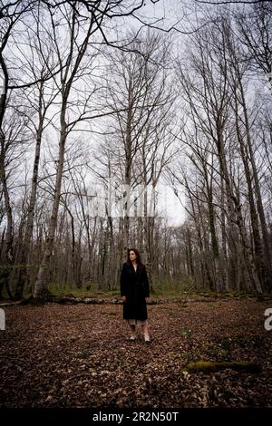 Solo una giovane donna che cammina attraverso una foresta di faggi con una coperta di foglie a terra vestita con un lungo cappotto nero. Concetto mistico in natura Foto Stock