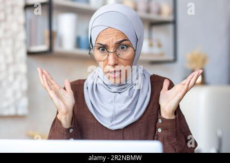Primo piano. Una giovane donna musulmana in un hijab e gli occhiali si siedono a casa di fronte a un notebook e guarda lo schermo in shock, le spalma le mani. Foto Stock