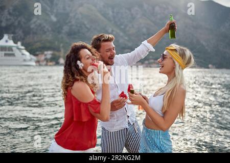 Amici felici che si godono una giornata al lago ridendo e mangiando anguria e bevendo birre. Foto Stock