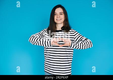 Joyful Woman in Striped Sweater sorridente, gestante mani del cuore - simbolo di amore e felicità - isolato su sfondo blu - perfetto per positivo Foto Stock