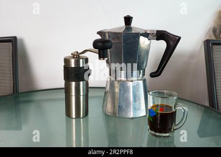 Macinacaffè Hario, moka pot, e tazza di vetro con caffè sedersi su un tavolo di vetro davanti ad una parete bianca/ Foto Stock