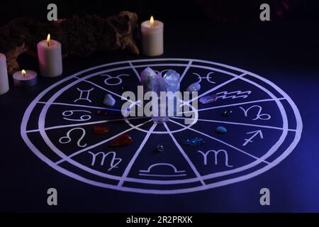 Pietre naturali per i segni zodiacali, grafico astrologico disegnato e candele brucianti su tavola blu scuro Foto Stock