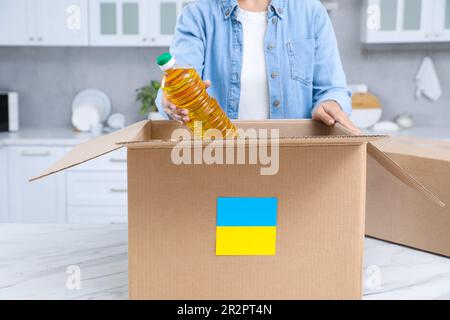 Aiuto umanitario a favore dell'Ucraina. Donna che mette una bottiglia di olio in una scatola di donazione al tavolo bianco al coperto, primo piano Foto Stock