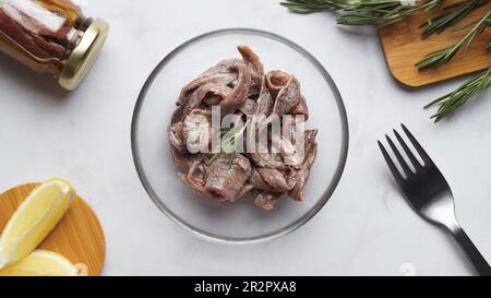 Filetti di acciuga in scatola di vetro, spicchi di rosmarino e limone su tavola di marmo bianco, piatto Foto Stock