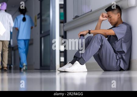 Un medico biraciale stanco che indossa scrub seduti nel corridoio dell'ospedale Foto Stock