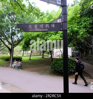 Cartello bilingue cinese inglese al Daan Park che indica il parco giochi con i visitatori seduti su panca e a piedi; Taipei, Taiwan. Foto Stock