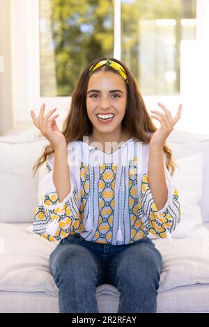 Biracial giovane donna allegra gesturing e guardando la macchina fotografica mentre si siede sul divano in soggiorno Foto Stock