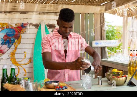 Barista afro-americano con shaker e preparazione di cocktail al bar sulla spiaggia, inalterato Foto Stock