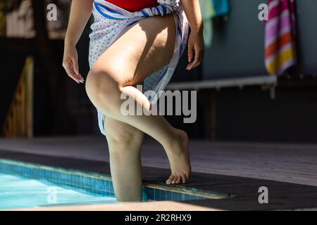 Parte bassa di donna biraciale inalterata in costume da bagno rosso e asciugamano che entra in piscina. Estate, tempo libero, relax e vacanze. Foto Stock