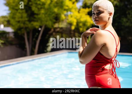 Donna biraciale alla moda inalterata in costume da bagno rosso con piscina in giardino soleggiato, spazio copia. Estate, tempo libero, relax e vacanze. Foto Stock