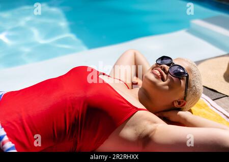 Felice donna biraciale alla moda inalterata in occhiali da sole e costume da bagno prendere il sole in piscina, sorridendo. Estate, tempo libero, relax e vacanze. Foto Stock