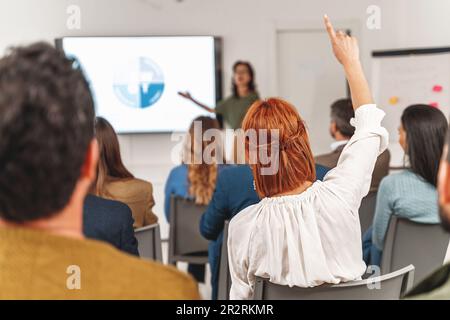 Donna d'affari con capelli rossi che alzano la mano per fare domanda durante una presentazione di analisi SWOT in grande stanza dell'ufficio. Pubblico diversificato, visto dal retro. Foto Stock