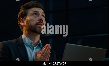 Speranzoso sognando uomo d'affari caucasico di mezza età uomo adulto seduto con il computer alla sera ufficio buio chiedere a dio benedire aiuto pregare speranza dire preghiere Foto Stock