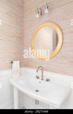 Un bagno con carta da parati marrone, un lavandino a piedistallo bianco, specchio circolare dorato, lavagna bianca e pareti a listelli. Foto Stock