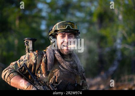 Moderno soldato mercenario fumante. Un uomo di mezza età barbuto in munizioni militari fuma un sigaro e guarda la telecamera. Primo piano verticale. Foto Stock