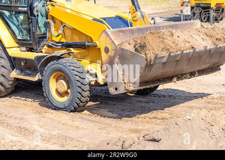 Lavori di scavo per il livellamento del cantiere per la costruzione di strade, un trattore con una benna grande trasporta il terreno. Foto Stock