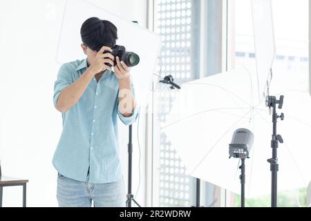 Un fotografo maschile asiatico in piedi e sparare nel suo studio. Con luci da studio come sfondo e spazio copia. Foto Stock