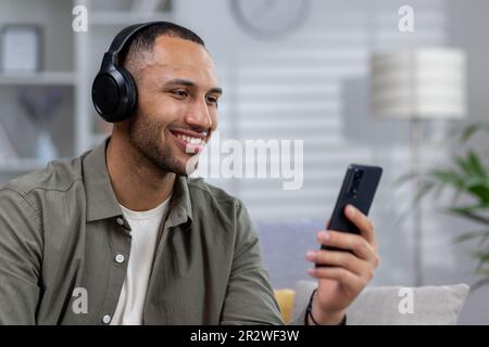 Giovane uomo ispanico seduto a casa con cuffie, tenendo il telefono in mano e ascoltando musica, audiolibro, podcast, parlando in videochiamata. Chiudi- Foto Stock