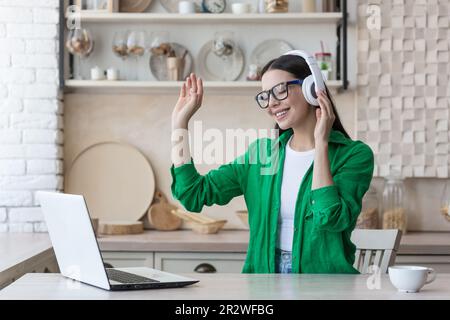 Una giovane e bella studentessa è seduta a casa in cucina utilizzando un computer portatile e indossando le cuffie. Ascolta musica, danze, alzò le mani, chiuse gli occhi, riposa, si rilassa. Foto Stock