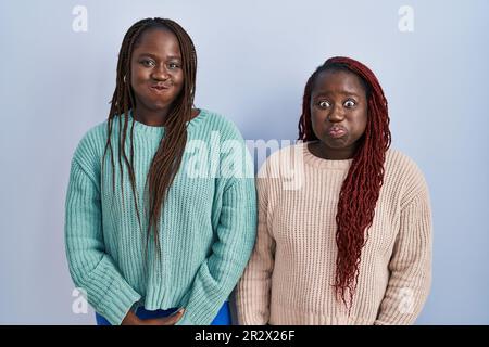 Due donne africane in piedi su sfondo blu puffing guance con faccia divertente. bocca gonfiata con aria, espressione pazza. Foto Stock