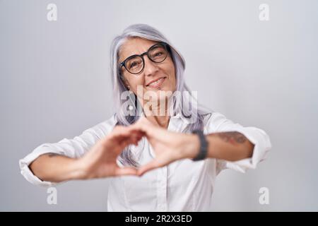 Donna di mezza età con tatuaggi che indossano gli occhiali in piedi su sfondo bianco sorridente nell'amore facendo forma simbolo del cuore con le mani. concetto romantico. Foto Stock