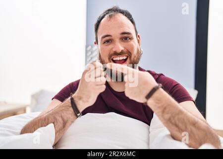 Più misura uomo ispanico con barba nel letto che tiene pillole sorridente felice indicando con mano e dito Foto Stock